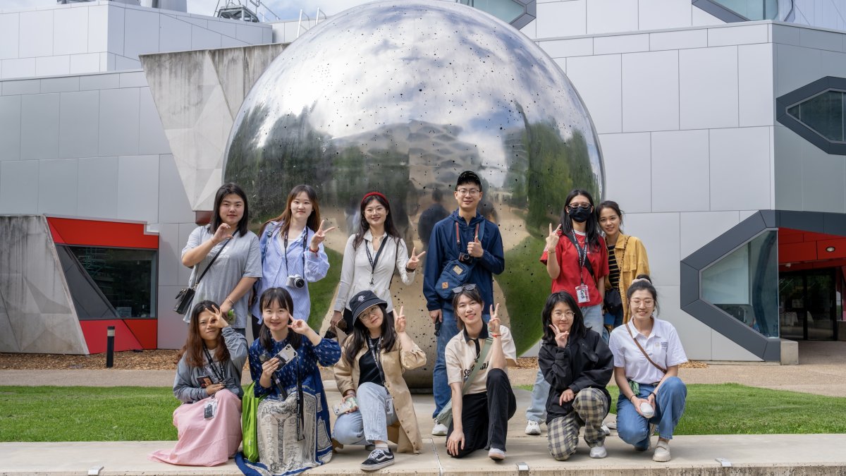 A group of people smiling with a large silver ball sculpture behind them. 