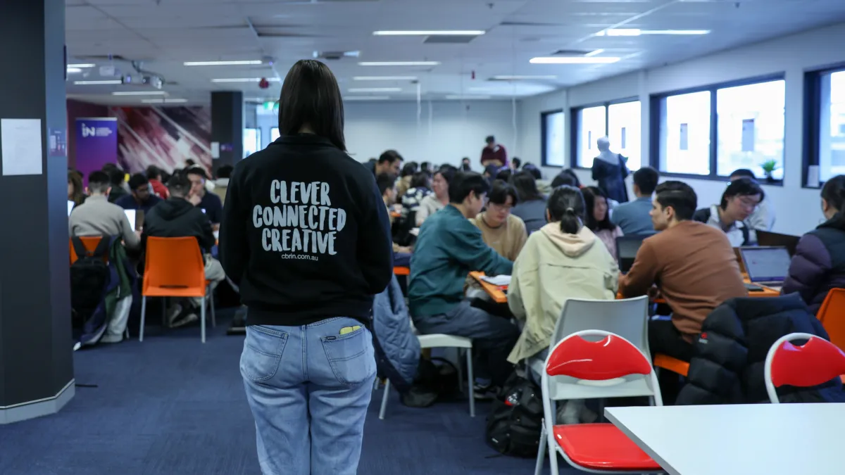 A woman standing with her back to the camera looking at a group of students during a Hackathon. 