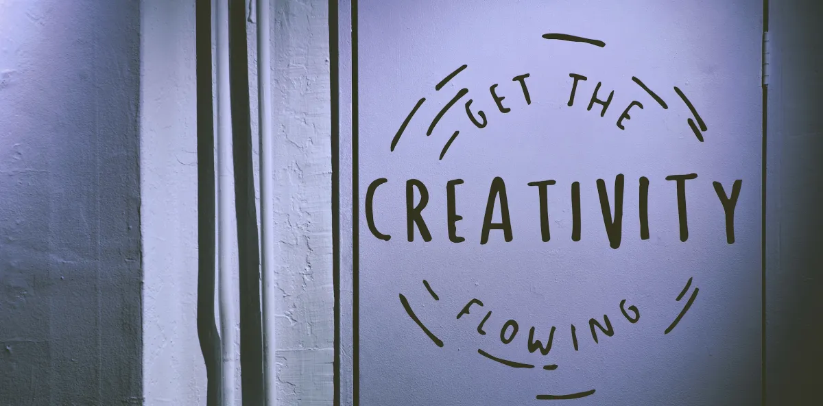 A white wall with two pipes vertically hanging on the left side with text on the right side inside a broken circle reads, &quot;Get the creativity flowing&quot;