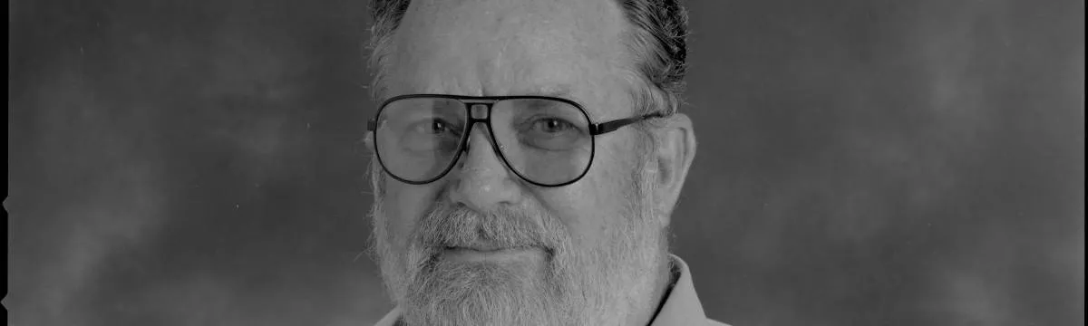 Gray scaled headshot of a mean with a white beard and large glasses.