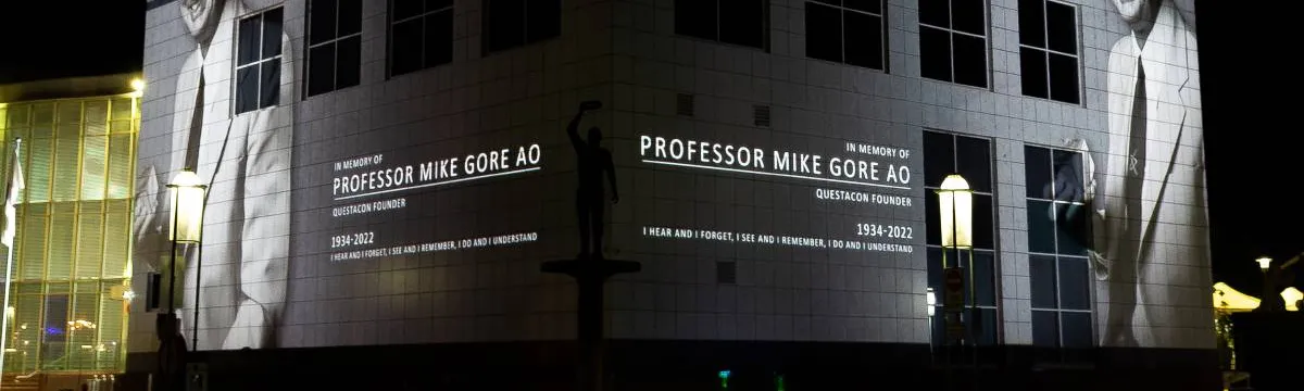 Projected image of a man in a suit and text that reads, Professor Mike Gore AO, on a building at night.