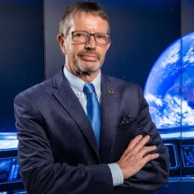 Man in a suit with hands crossed an a graphic display of planet earth in the background.
