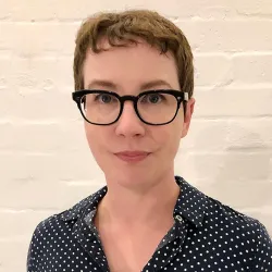 Headshot of a woman in glass in front of a white brick wall.