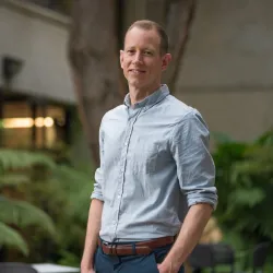 Headshot of a man with larger ferns and a building behind him.