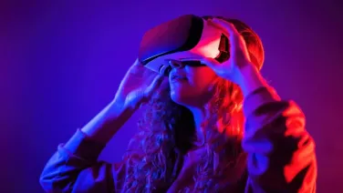 Woman holding a Virtual Augmented Reality goggles to her face. Background is a dark purple with a red light glow on the back of the woman.