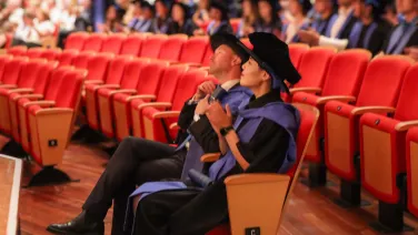 A man and a woman in graduation gowns and their diplomas in hand, clapping for those crossing the stage to get their diploma.