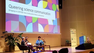 Three people sitting in chairs as panelist with a large projector behind them with the words, "Queering in science communication: Brining an LGBTQIA+ lens to #scicomm theory and practice.