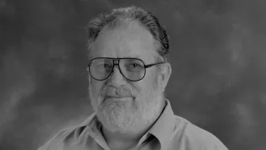 Gray scaled headshot of a mean with a white beard and large glasses.